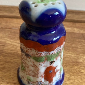 May include: A blue and white ceramic salt shaker with a floral design and a woman in a red kimono. The shaker has a round top with holes for dispensing salt.