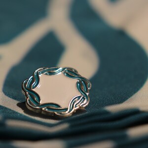 May include: A silver and teal enamel pin with a white center. The pin is shaped like a circle with a decorative border.