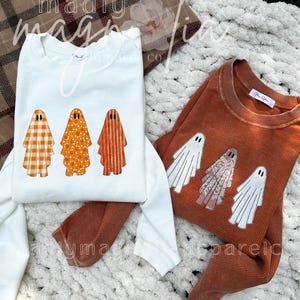 May include: Two long-sleeved shirts, one white and one rust-coloured, with ghost graphics. The white shirt features three ghosts with different patterns, while the rust shirt has three white ghost designs. The shirts are displayed on a white, textured surface.