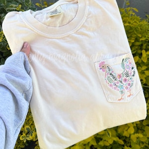 Op de afbeelding: Een licht beige t-shirt met een geborduurd bloemenpatroon van een kip op de zak. Het shirt heeft een ronde hals en korte mouwen.