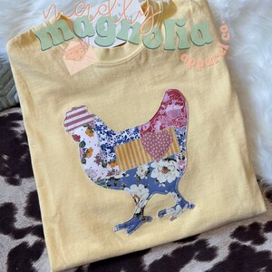 May include: A yellow t-shirt with a patchwork-style chicken design. The design is made up of various floral and geometric patterns in shades of blue, pink, red, and yellow. The text "merrily magnolia apparel co." is visible in the top right corner of the image.