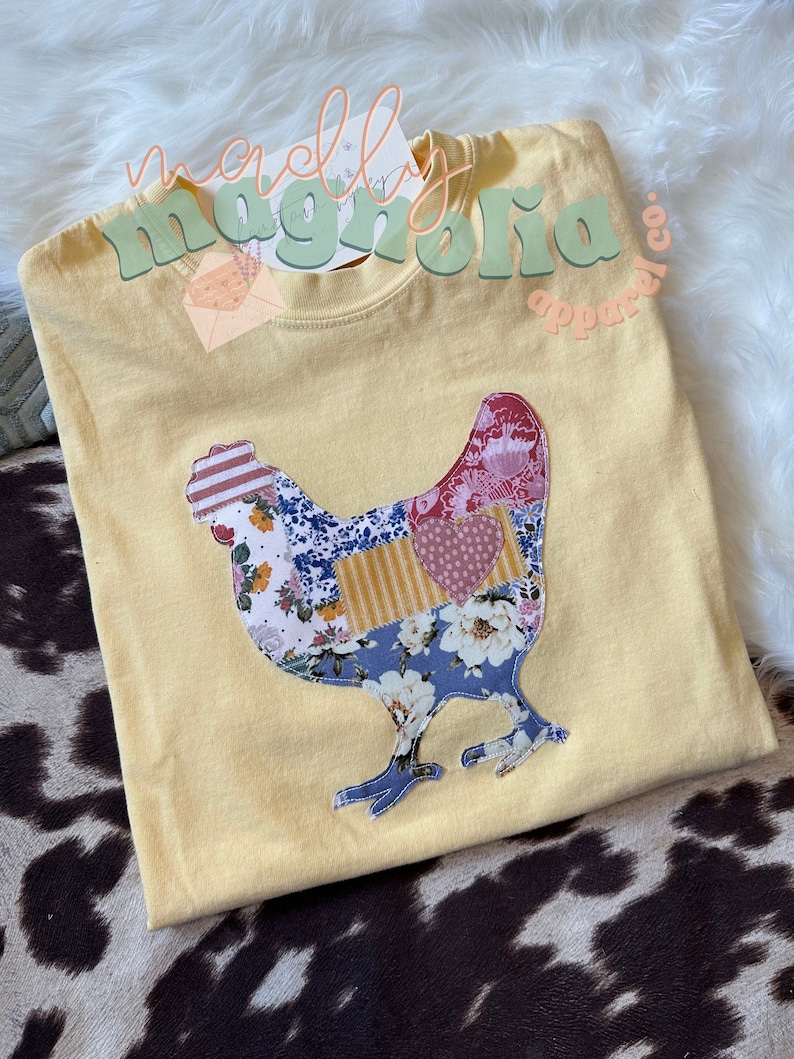 May include: A yellow t-shirt with a patchwork-style chicken design. The design is made up of various floral and geometric patterns in shades of blue, pink, red, and yellow. The text "merrily magnolia apparel co." is visible in the top right corner of the image.