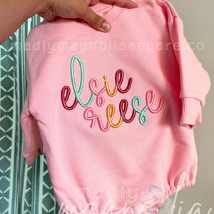 Könnte beinhalten: Ein hellrosa Langarm-Sweatshirt-Strampler mit dem gestickten Namen Elsie Reese in Schreibschrift. Die Buchstaben sind in abwechselnden Farben in Türkis, Dunkelrosa und Orange gehalten. Das Kleidungsstück hat einen gerippten Rundhalsausschnitt.