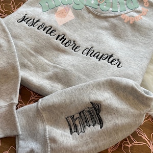 May include: A gray sweatshirt with the text "just one more chapter" embroidered on the front. The sweatshirt has a black and white graphic on the sleeve.
