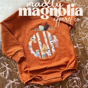 May include: Orange baby onesie with a floral monogram in the shape of a pumpkin. The onesie has long sleeves and snaps at the bottom. The text "madly magnolia apparel co." is printed on the onesie.