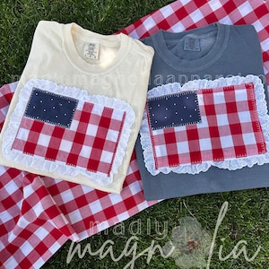 May include: Two t-shirts, one cream and one gray, each adorned with a patriotic flag design. The flag features a navy blue field with white stars and red and white gingham stripes, framed by a white lace border. The shirts rest on a red and white checkered picnic blanket.