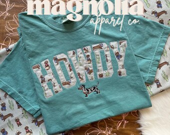 Può includere: Una t-shirt a maniche corte color verde acqua con la scritta HOWDY in stampatello e un motivo di bassotti e cactus. Sotto il testo è presente un bassotto maculato ricamato. La maglietta è appoggiata su un tessuto con motivo a cani.
