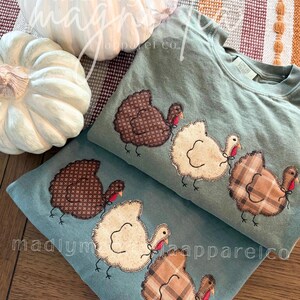 Op de afbeelding: Twee saliegroene sweatshirts met ronde hals en een applicatie van drie kalkoenen. De kalkoenen zijn gemaakt van bruine stof met patroon, cr&egrave;me en beige ruitjesstof met gestikte contouren, een rode lel en zwarte geborduurde pootjes.