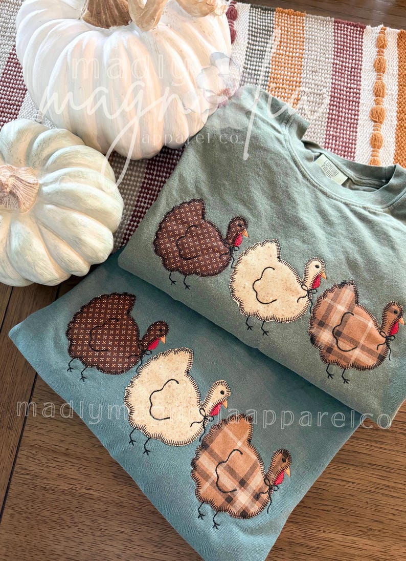 Op de afbeelding: Twee saliegroene sweatshirts met ronde hals en een applicatie van drie kalkoenen. De kalkoenen zijn gemaakt van bruine stof met patroon, cr&egrave;me en beige ruitjesstof met gestikte contouren, een rode lel en zwarte geborduurde pootjes.