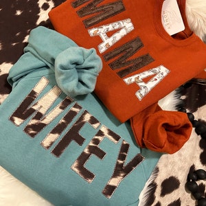 May include: Two sweatshirts with the words "Mama" and "Wifey" in brown faux leather letters. The "Mama" sweatshirt is rust colored and the "Wifey" sweatshirt is teal.