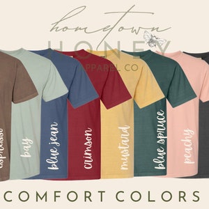May include: A row of eight t-shirts in different colors, including espresso, bay, blue jean, crimson, mustard, blue spruce, peachy, pepper, and ivory. The shirts are all the same style and have the text "COMFORT COLORS" printed below them.