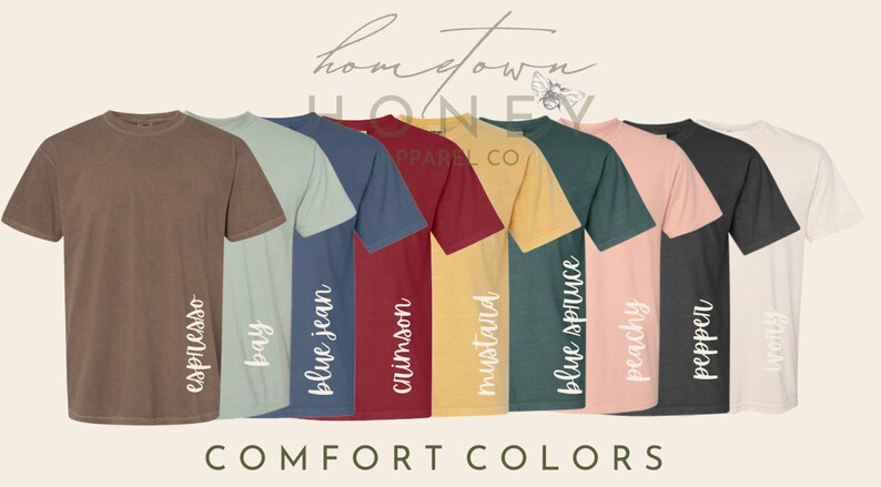 May include: A row of eight t-shirts in different colors, including espresso, bay, blue jean, crimson, mustard, blue spruce, peachy, pepper, and ivory. The shirts are all the same style and have the text "COMFORT COLORS" printed below them.