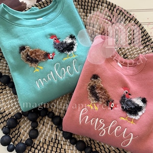 Op de afbeelding: Twee crewneck sweatshirts, één in teal en één in roze, elk versierd met geborduurde kippen. Het teal sweatshirt heeft de naam "mabel", de roze "hazley". Beide liggen op een gevlochten mand.