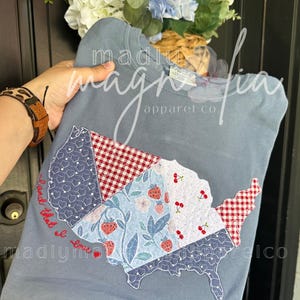May include: A blue-grey sweatshirt featuring a patchwork design of the United States. The patchwork includes patterns of hearts, strawberries, and gingham. The text "Land that I love" is embroidered on the shirt. The brand name "madly magnolia apparel co." is also visible.