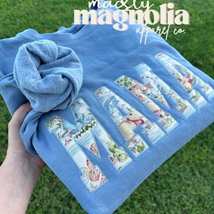 Może przedstawiać: Jasno błękitna bluza z napisem "MAMA" napisanym dużymi, kolorowymi literami. Litery są wykonane z tkaniny z kwiatowym wzorem i są przyszyte do bluzy. Tekst "madly magnolia apparel co." jest nadrukowany na g&oacute;rze bluzy na biało.