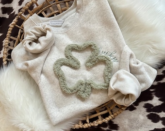 May include: A light beige sweatshirt with a green textured shamrock design and the word "lucky" embroidered in green. The sweatshirt is folded in a woven basket on a white fluffy surface, set against a cowhide pattern.