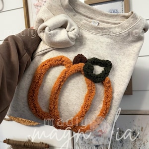 Könnte beinhalten: Ein hellbeige Sweatshirt mit einem strukturierten orangefarbenen Kürbismotiv und einem grünen Stiel. Der Kürbis besteht aus einem weichen, geschlungenen Stoff. Dies ist ein Bekleidungsartikel zum Thema Herbst.