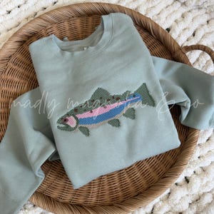 May include: A sage green crewneck sweatshirt featuring a cross-stitch style embroidered rainbow trout graphic on the center chest. The fish design incorporates shades of pink, blue, and dark green thread, creating a textured appearance.