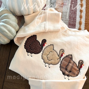 Op de afbeelding: Een cr&egrave;mekleurig sweatshirt met lange mouwen en drie geappliqueerde kalkoenen op de voorkant. De kalkoenen zijn gemaakt van verschillende stoffen: &eacute;&eacute;n donkerbruin met een klein patroon, &eacute;&eacute;n beige en &eacute;&eacute;n bruin geruit. Elke kalkoen heeft een rode lel.