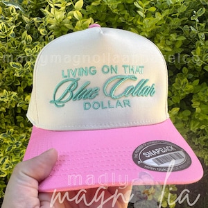 Op de afbeelding: Een witte en roze snapback pet met de tekst "LIVING ON THAT Blue Collar DOLLAR" in turquoise geborduurd. De pet heeft een roze klep en een "SNAPBACK" label.