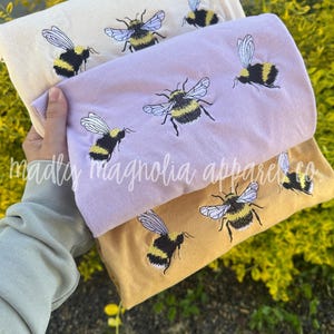 May include: Three folded sweatshirts in cream, lavender, and mustard yellow, each embroidered with multiple bumblebees. The text "madly magnolia apparel co" is visible on the bottom left of the image.