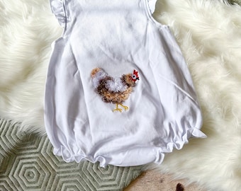 May include: White baby onesie with a ruffled neckline and short sleeves. The onesie has a brown and white embroidered chicken on the back.