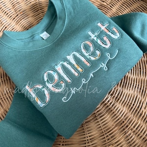 May include: A dark teal long-sleeve sweatshirt featuring the name Bennett appliqued in a floral patterned fabric with white stitching. The sweatshirt has a crew neckline and a soft fleece interior, showcasing a custom embroidered design.