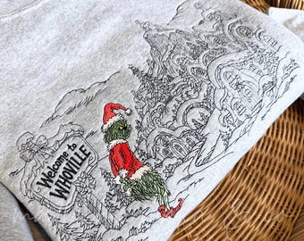Może przedstawiać: Jasnoszara bluza z haftem przedstawiającym zieloną postać w czerwonym stroju Świętego Mikołaja obok znaku z napisem Welcome to WHOVILLE oraz szczegółowym czarnym konturem zaśnieżonej wioski.