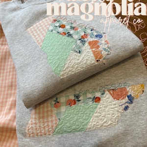 May include: Two gray sweatshirts with a Tennessee state design. The design is a patchwork of patterned fabrics, including floral, gingham, and solid colors, stitched in the shape of the state. The text "Tennessee" is embroidered on the design.