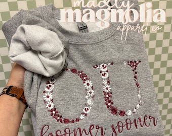May include: Gray sweatshirt with a floral embroidered "OU" and the text "Boomer Sooner" in maroon. The sweatshirt is folded and held by a hand. The text "madly magnolia apparel co." is visible in the top right corner.