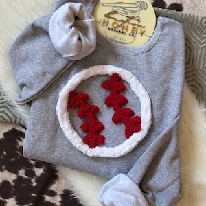 May include: Gray sweatshirt with a white and red baseball design. The baseball is made of fluffy material. The sweatshirt has the text "Honey Apparel Co. Est 2020" on the front.