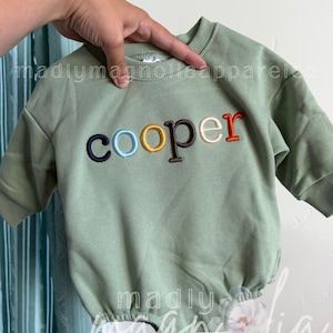 Könnte beinhalten: Ein langärmeliger Sweatshirt-Strampler in Salbeigrün mit dem Namen cooper, der in mehrfarbigen Buchstaben auf der Brust gestickt ist, darunter Marineblau, Hellblau, Gelb, Braun, Creme und Orange, mit geripptem Rundhalsausschnitt und Bündchen.