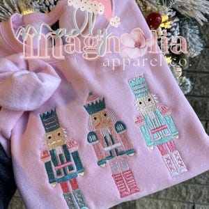 May include: Pink sweatshirt with embroidered nutcracker designs. The text "Madly Magnolia Apparel Co." is on the front of the sweatshirt.