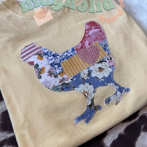 May include: A yellow t-shirt with a patchwork chicken design. The chicken is made of different fabrics with floral and geometric patterns. The text "maclaly magnolia apparel co." is printed on the shirt.