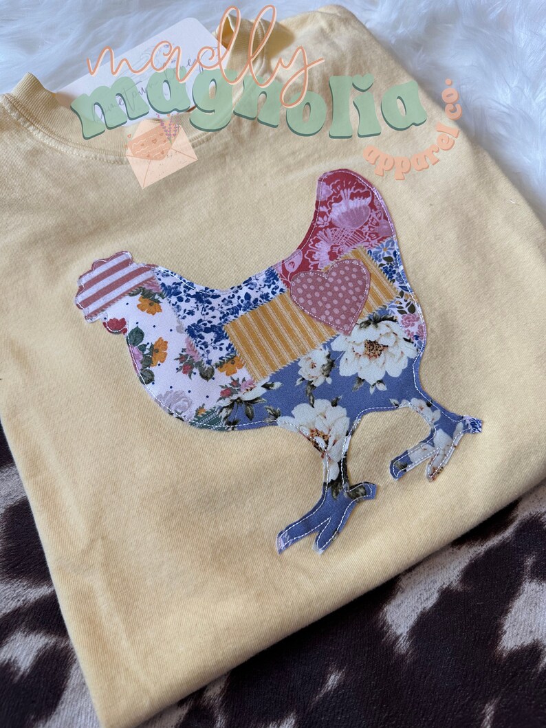 May include: A yellow t-shirt with a patchwork chicken design. The chicken is made of different fabrics with floral and geometric patterns. The text "maclaly magnolia apparel co." is printed on the shirt.