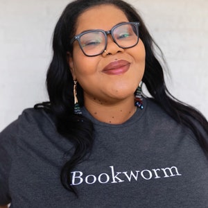 May include: A woman wearing black framed glasses and a dark gray t-shirt with the word "Bookworm" embroidered in white on the front.