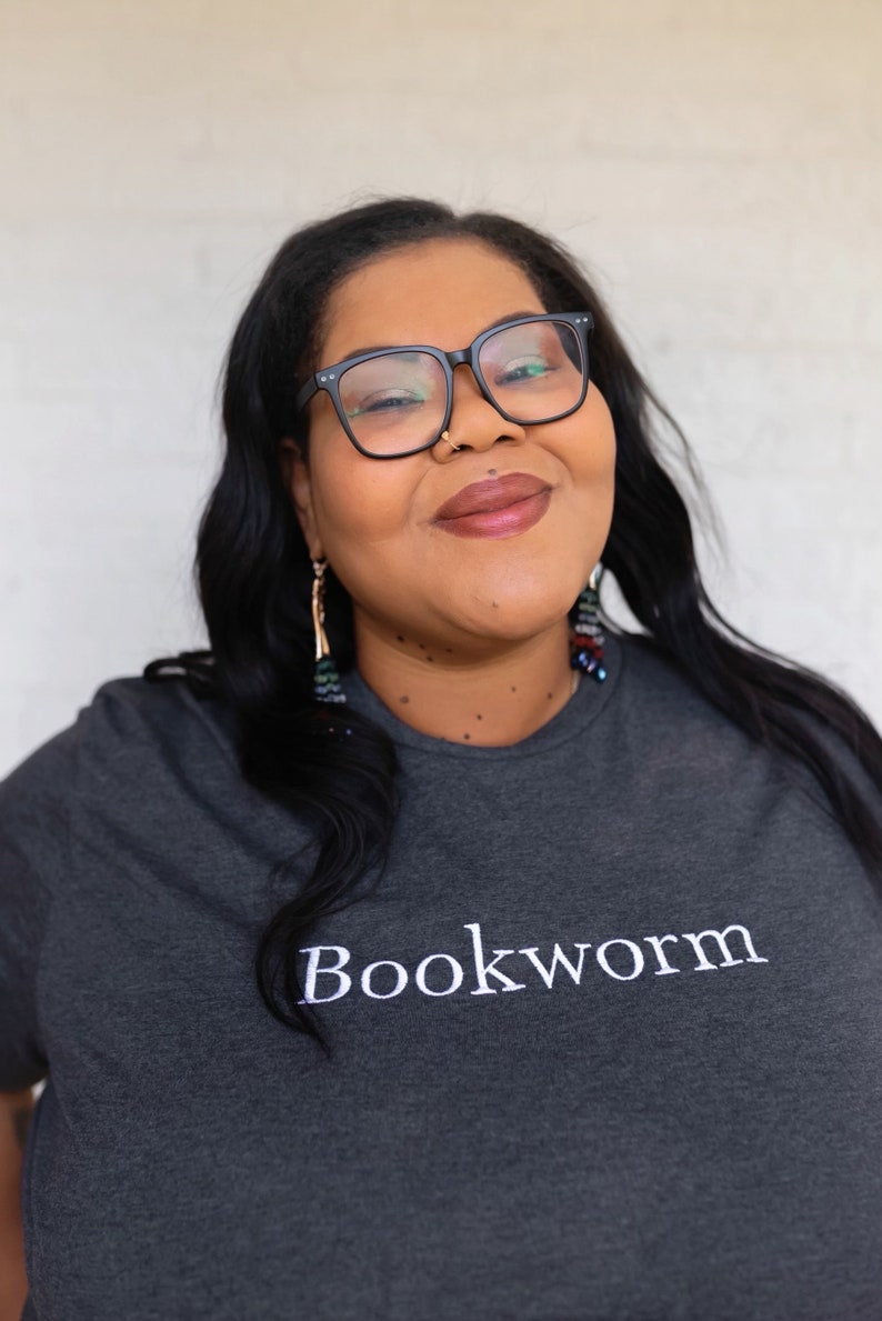 May include: A woman wearing black framed glasses and a dark gray t-shirt with the word "Bookworm" embroidered in white on the front.