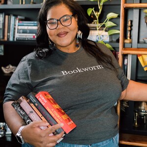 May include: A woman wearing a gray t-shirt with the word "Bookworm" printed on it. She is holding a stack of five books in her left hand. The books are all paperback and have colorful covers. The woman is standing in front of a bookshelf.