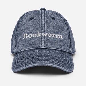 Puede incluir: Una gorra de b&eacute;isbol de mezclilla azul con la palabra "Bookworm" bordada en blanco en la parte delantera.
