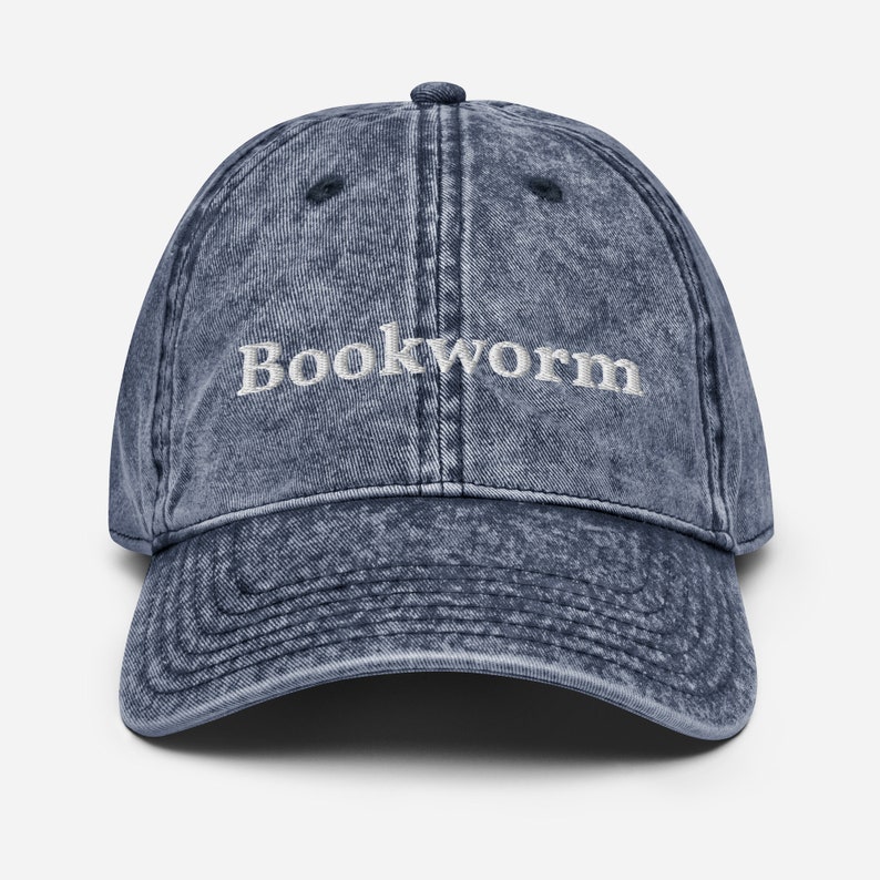 Puede incluir: Una gorra de b&eacute;isbol de mezclilla azul con la palabra "Bookworm" bordada en blanco en la parte delantera.