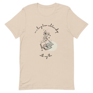 May include: A beige t-shirt with a black graphic design of a stack of books with a flower growing from the top. The text "in a long-term relationship with my thr" is written below the graphic.