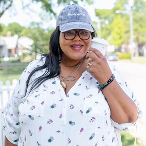 Puede incluir: Una mujer con una camisa blanca de botones con estampado de pi&ntilde;a, una gorra de b&eacute;isbol de mezclilla gris con el texto "Bookworm" bordado en la parte delantera y una pulsera de cuentas negras. Est&aacute; sosteniendo una taza de caf&eacute; para llevar blanca.