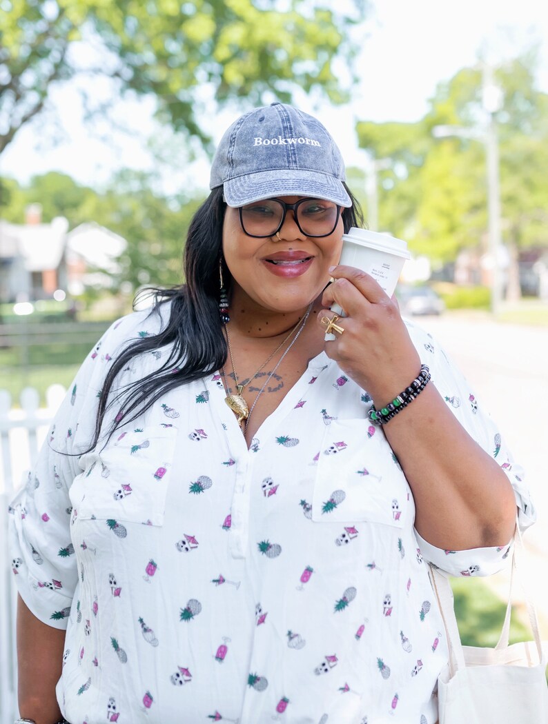 Puede incluir: Una mujer con una camisa blanca de botones con estampado de pi&ntilde;a, una gorra de b&eacute;isbol de mezclilla gris con el texto "Bookworm" bordado en la parte delantera y una pulsera de cuentas negras. Est&aacute; sosteniendo una taza de caf&eacute; para llevar blanca.