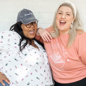 Puede incluir: Dos mujeres est&aacute;n riendo juntas. Una mujer lleva una camisa blanca con estampado de pi&ntilde;a y una gorra de b&eacute;isbol negra con la palabra "Bookworm" bordada. La otra mujer lleva una sudadera de color melocot&oacute;n con las palabras "Romance Genre" impresas.