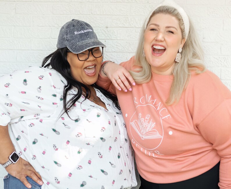 Puede incluir: Dos mujeres est&aacute;n riendo juntas. Una mujer lleva una camisa blanca con estampado de pi&ntilde;a y una gorra de b&eacute;isbol negra con la palabra "Bookworm" bordada. La otra mujer lleva una sudadera de color melocot&oacute;n con las palabras "Romance Genre" impresas.