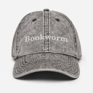 Puede incluir: Una gorra de b&eacute;isbol de estilo vintage gris desgastada con la palabra "Bookworm" bordada en blanco en la parte delantera.