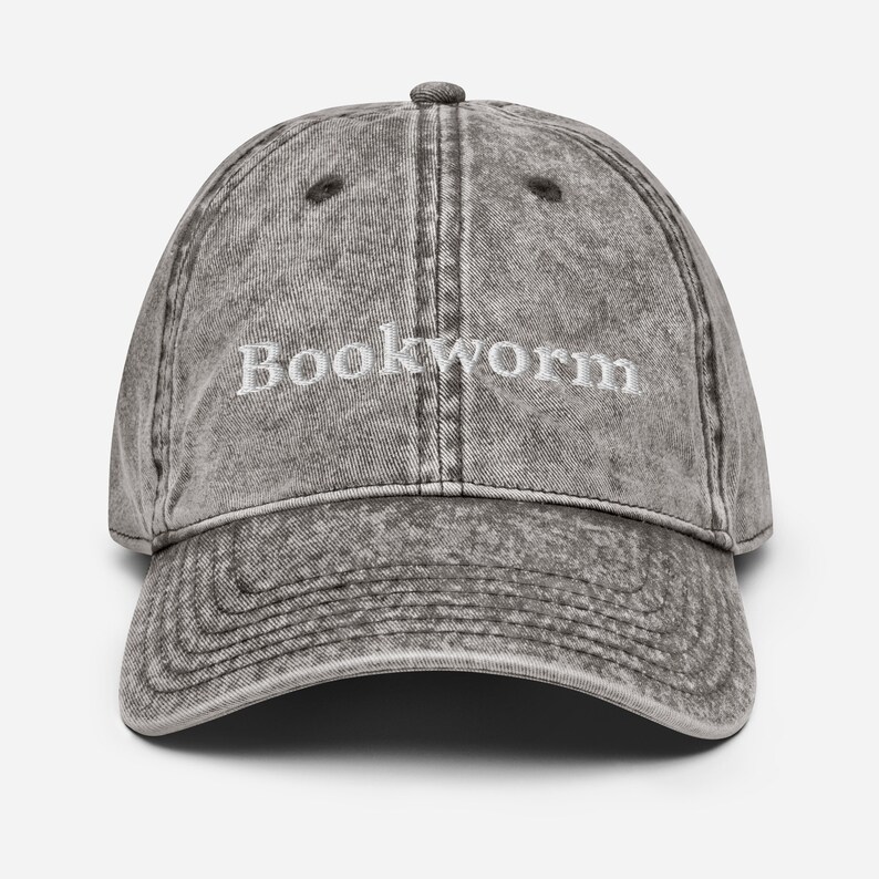 Puede incluir: Una gorra de b&eacute;isbol de estilo vintage gris desgastada con la palabra "Bookworm" bordada en blanco en la parte delantera.