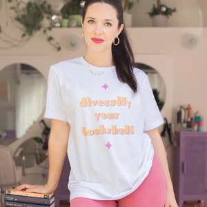 May include: A woman wearing a white t-shirt with the text "diversify your bookshelf" in orange and pink letters. She is wearing pink shorts and is holding a stack of books. The books have titles such as "Trouble the Saints", "Woman of Light", "Off Her Head", and "Caste".