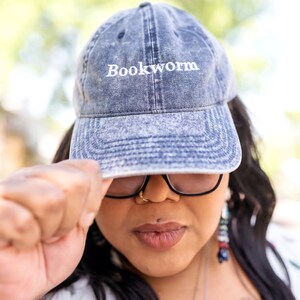Puede incluir: Una mujer con una gorra de b&eacute;isbol de mezclilla azul con la palabra "Bookworm" bordada en blanco. Tambi&eacute;n lleva gafas y tiene un piercing en la nariz.