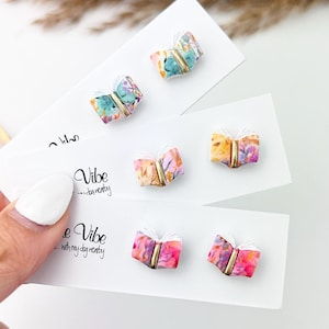 May include: A hand holds several white cards with colorful book-shaped earrings. The earrings have a gold spine and pages with a watercolor design in shades of pink, blue, yellow, and green. The cards have the word "Vibe" printed on them.